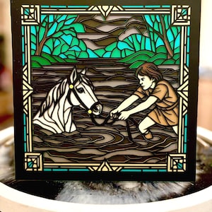 May include: A layered art piece depicting a person in a brown tunic leading a white horse through water. The artwork features a black border with teal and gold accents, and a background of trees and water.
