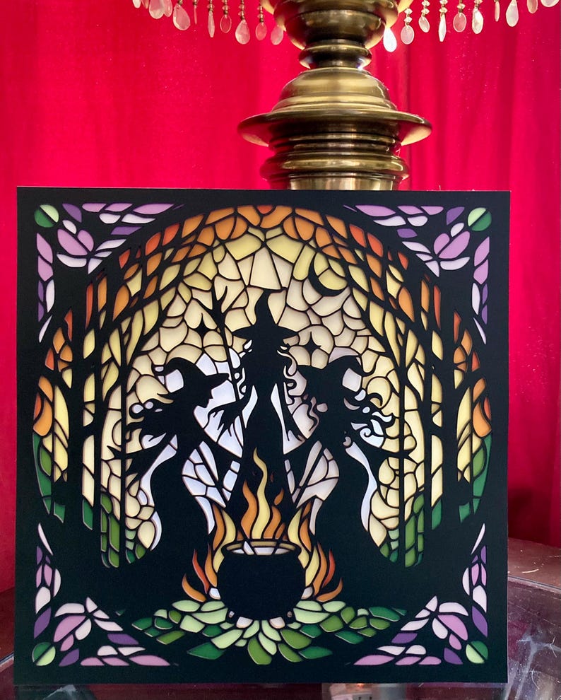 May include: A black paper cut-out of three witches standing around a cauldron with flames. The witches are silhouetted against a stained glass background with a sunset and forest scene.