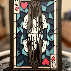 Puede incluir: Una pieza de arte en capas que se asemeja a una carta de juego, con una figura de pelo largo negro y una camisa blanca. La carta está enmarcada en negro, con los símbolos de la Reina de Corazones en las esquinas, sobre un fondo de estilo vidriera.