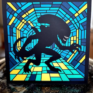 May include: Black silhouette of an Alien creature against a stained-glass style background. The background features a geometric pattern in shades of blue, yellow, and green. The artwork is framed in black, creating a striking contrast.