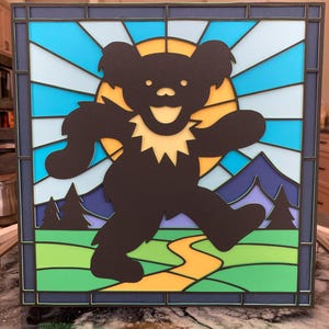 Puede incluir: Obra de arte enmarcada con la silueta de un oso negro sonriente, sobre un fondo de sol amarillo, cielo azul y paisaje verde. El diseño recuerda a una vidriera, con un borde azul oscuro.