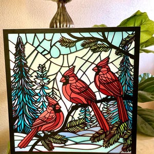 May include: A square, layered art piece featuring three red cardinal birds perched on a branch. The background depicts a winter scene with evergreen trees and a stained-glass effect, framed in black.