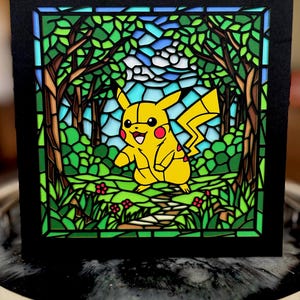 May include: A stained glass-style artwork featuring a yellow Pikachu character in a forest scene. The artwork has a black border and depicts trees, grass, and a blue sky. Red flowers and a path are also visible.