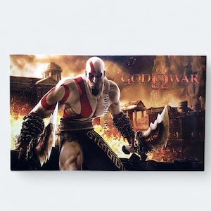 Peut inclure: Une jaquette de jeu vidéo représentant un personnage musclé à la peau rouge tenant une arme. Le personnage se tient devant un fond enflammé avec le texte "God of War 2" et "PlayStation 2" visible.