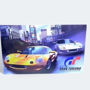 Peut inclure: Une voiture de sport jaune et argentée fonçant dans une rue de ville. La voiture est au premier plan de l'image, avec un horizon urbain en arrière-plan. L'image porte le texte "Gran Turismo The Real Driving Simulator" en bas.