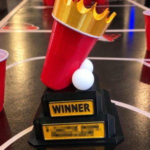 Könnte beinhalten: Eine Trophäe bestehend aus einem roten Plastikbecher mit einer goldenen Krone, montiert auf einem schwarzen, gestuften Sockel. Der Sockel zeigt ein gelbes Schild mit dem Wort WINNER in schwarzer Schrift sowie ein zweites gelbes Schild darunter.