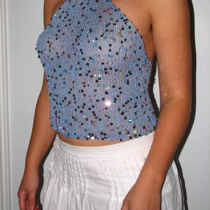 May include: A light blue halter top with a knitted texture, embellished with colorful sequins. The top has a cropped design and a halter neck. The garment is paired with a white skirt.