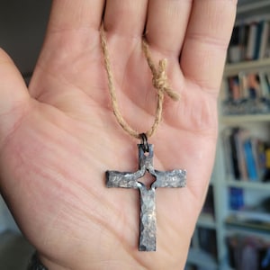 Può includere: Ciondolo a croce argentato fatto a mano con superficie strutturata e un ritaglio a forma di stella al centro. La croce è sospesa a un cordino in fibra naturale, annodato in alto.