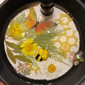 May include: A decorative bottle stopper with a black handle. The stopper features a butterfly, yellow flowers, a bee, and other floral elements encased in clear resin. The stopper is set in a black container.