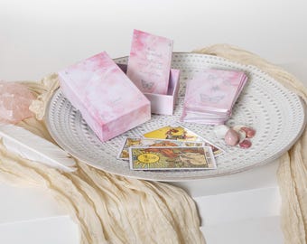 May include: A pink and white tarot card deck with a butterfly design, displayed on a white decorative plate with other items. The deck is accompanied by a box, cards, crystals, and a feather.