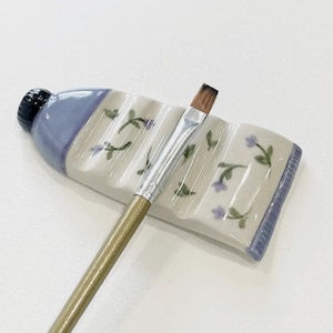 May include: A ceramic paint tube-shaped brush holder with a blue and white floral design. A paintbrush with a gold handle rests in the holder. The holder is designed to resemble a tube of paint.
