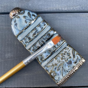 May include: A blue ceramic paint brush holder with floral and leaf patterns. The holder has a rectangular shape with a rounded top and a silver-colored rim. A paint brush with an orange bristle is resting on the holder.