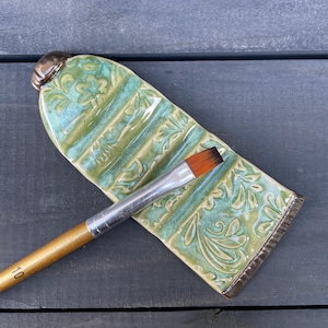 Könnte beinhalten: Keramik-Palette in Form einer Farbtube, mit grünem und blauem Blumenmuster. Ein Pinsel mit Holzgriff und der Größenangabe 10 liegt auf der Palette. Die Palette ist etwa 15 cm lang.