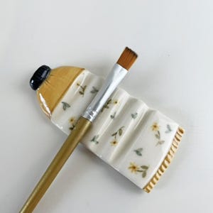 May include: A white ceramic paintbrush holder shaped like a bottle with a floral design. The holder has a yellow rim and a black cap. A gold paintbrush is resting on the holder.