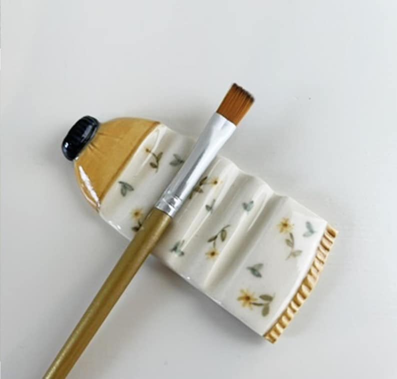 May include: A white ceramic paintbrush holder shaped like a bottle with a floral design. The holder has a yellow rim and a black cap. A gold paintbrush is resting on the holder.