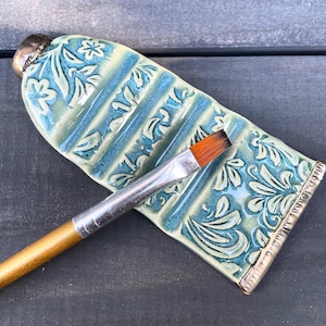 Könnte beinhalten: Eine Keramik-Farbpalette in Form einer Tube, mit einem floralen Design in Blau und Weiß. Ein Pinsel mit silberner Zwinge und Holzgriff liegt auf der Palette. Die Palette ist etwa 15 cm lang.