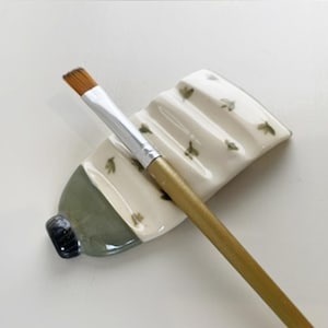 Può includere: Un portapennelli in ceramica a forma di tubetto di vernice con un pennello piatto e angolato all'interno. Il supporto è bianco sporco con accenti verdi e piccoli disegni di foglie verdi. Il pennello ha un manico dorato e una ghiera argentata.