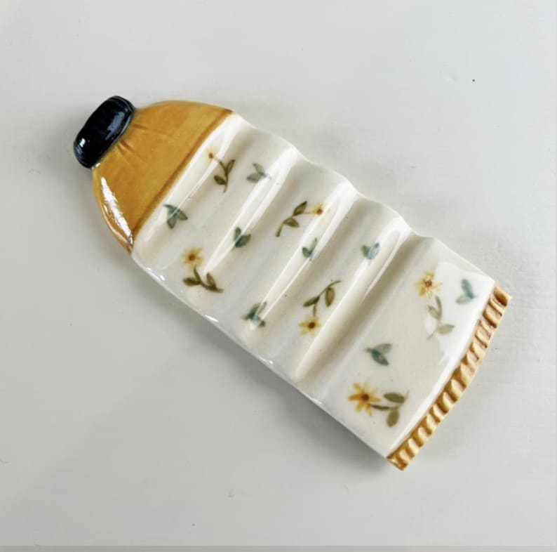May include: A ceramic dish shaped like a bottle with a yellow and white floral design. The dish has five ridges and is perfect for holding chopsticks or utensils.