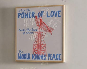 Może przedstawiać: Kwadratowy wydruk ścienny w jasnej drewnianej ramie z czerwoną grafiką liniową wieży radiowej i niebieskim tekstem: When the POWER OF LOVE beats the love of power the WORLD KNOWS PEACE. Wydruk jest umieszczony na ścianie.
