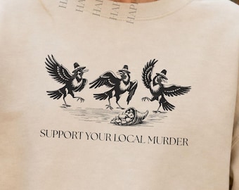 May include: A sand-colored crewneck sweatshirt featuring a black graphic of three crows and a cornucopia with the text "SUPPORT YOUR LOCAL MURDER" printed below. The garment is a Gildan crewneck sweater in size XL, as indicated by the visible labels.