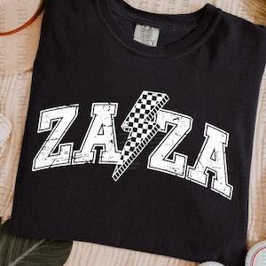 May include: Black t-shirt with the word "ZAZA" in distressed white lettering and a checkered lightning bolt graphic. The shirt is a comfortable, casual style.