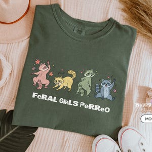 May include: Moss green t-shirt featuring a playful illustration of four raccoons in various poses. The text "FeRAL GiRLS PeRReO" is printed below the raccoons. The shirt is paired with white sneakers and a beige hat.