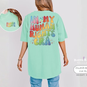 May include: A light green t-shirt with the text "IN MY HUMAN RIGHTS ERA" in colorful, retro-style lettering on the back. The front of the shirt has the text "HUMAN RIGHTS" in rainbow colors. The shirt is size XL.