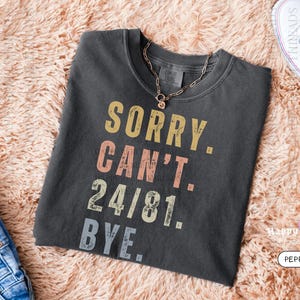 May include: Dark gray t-shirt with the text "SORRY. CAN'T. 24/81. BYE." in a retro font. A gold necklace is draped on the shirt. The shirt is folded on a peach-colored, fluffy surface. White sneakers are visible in the background.