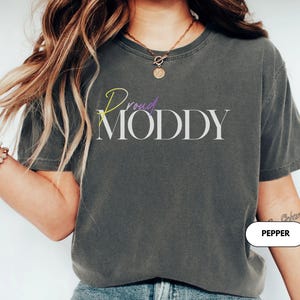 May include: Dark gray t-shirt with the text "Proud Moddy" in white, with the words "Proud" in yellow and purple. The shirt is a casual style, suitable for everyday wear.