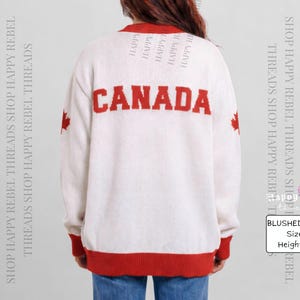 Może przedstawiać: Sweter z dzianiny w kolorze kości słoniowej z napisem CANADA wyhaftowanym pogrubionymi czerwonymi literami na plecach. Odzież posiada czerwone wykończenia przy kołnierzu, mankietach i u dołu oraz czerwony liść klonu na każdym rękawie. Widoczny jest tekst BLUSHED IVORY, Size L, Height: 5'3.