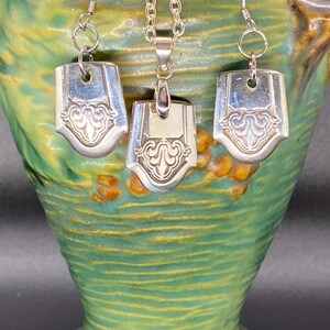 May include: A silver necklace and earrings set with a decorative design. The pendant and earrings are made from repurposed silverware.