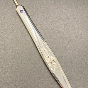 May include: A silver seam ripper with a decorative rose design. The tool has a pointed blade with a red tip and a purple gem on the handle. This sewing tool is used to remove stitches.
