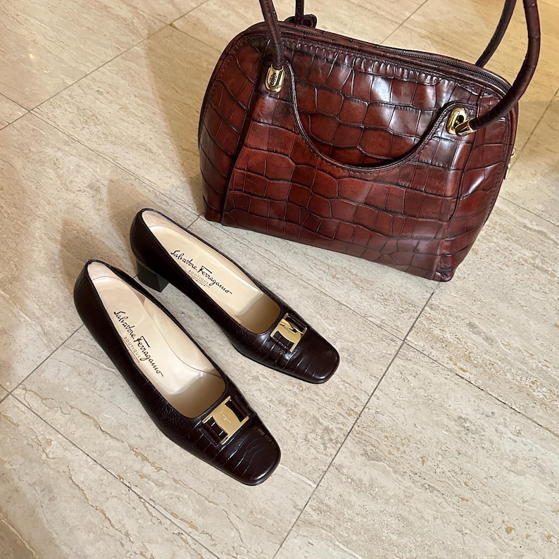 May include: A matching set of dark brown leather Salvatore Ferragamo shoes and handbag. The shoes have a square toe and a gold-tone buckle. The handbag has a crocodile-like texture and a top handle.