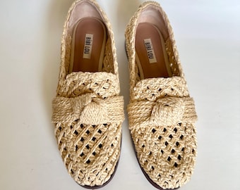 May include: A pair of beige woven loafers with a knotted detail on the vamp. The shoes have a lattice design and a brown sole. The brand name "BIBI LOU" is visible inside the shoe.