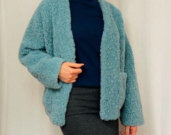 Puede incluir: Chaqueta azul claro con frente abierto y aspecto texturizado y esponjoso. La chaqueta tiene dos bolsillos delanteros y se usa sobre un cuello alto azul oscuro. El atuendo incluye una falda lápiz gris.