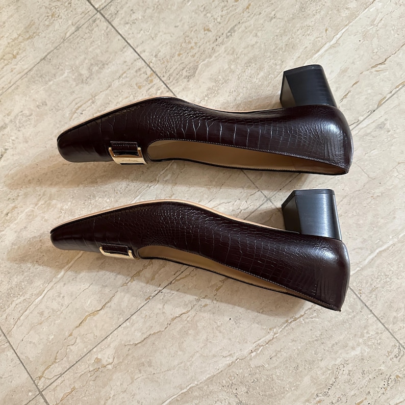May include: A pair of dark brown, textured leather pumps with a square toe and a small, block heel. The shoes have a gold-colored buckle detail and a neutral-colored interior.