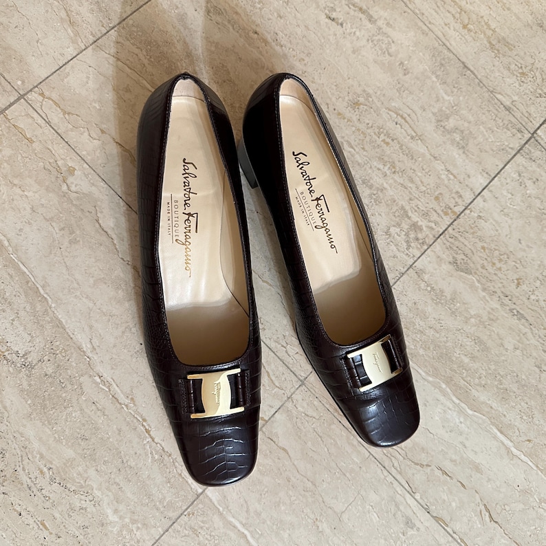 May include: A pair of dark brown leather Salvatore Ferragamo pumps. The shoes have a square toe, a low heel, and a gold-tone buckle. The interior of the shoe is a light beige color, and the brand name is printed in black.