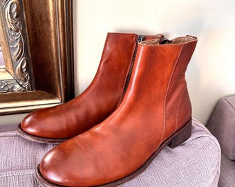May include: A pair of ankle boots crafted from smooth, cognac-colored leather. These boots feature a rounded toe, a low stacked heel, and a vertical zipper closure along the inner side. The leather has a polished finish with visible stitching.