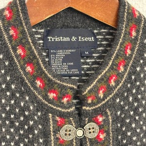 Puede incluir: Un cárdigan gris de Tristan & Iseut con un diseño floral en rojo, verde y crema. El cárdigan tiene cierre de botón y una etiqueta que dice "Tristan & Iseut". El suéter es talla M.