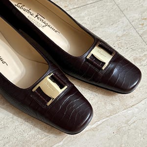 May include: A pair of dark brown leather Salvatore Ferragamo flats with a square toe and gold-tone buckle. The shoes have a textured, crocodile-like pattern and the brand name is visible inside the shoe.