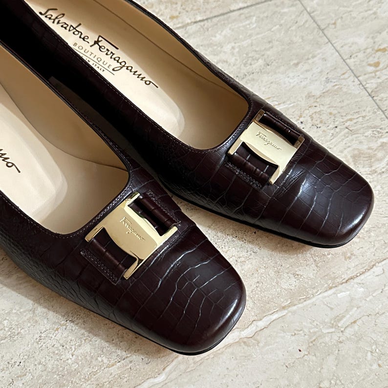 May include: A pair of dark brown leather Salvatore Ferragamo flats with a square toe and gold-tone buckle. The shoes have a textured, crocodile-like pattern and the brand name is visible inside the shoe.