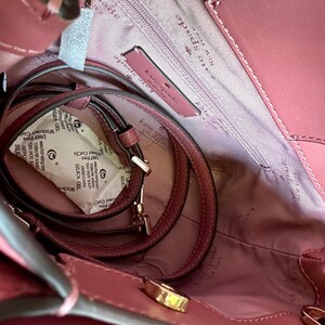 May include: A dusty rose-colored Kate Spade handbag, viewed from above, revealing the interior. The bag features a matching shoulder strap and gold-toned hardware. The interior lining is a lighter shade of pink, with the Kate Spade logo visible.
