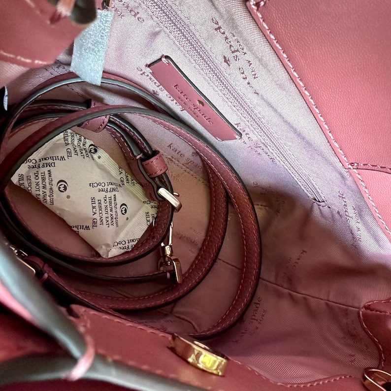 May include: A dusty rose-colored Kate Spade handbag, viewed from above, revealing the interior. The bag features a matching shoulder strap and gold-toned hardware. The interior lining is a lighter shade of pink, with the Kate Spade logo visible.