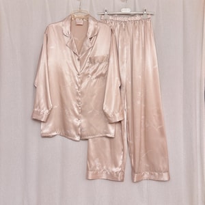 Puede incluir: Un conjunto de pijama de satén rosa claro, que incluye una camisa abotonada con bolsillo en el pecho y pantalones largos. El conjunto tiene un sutil estampado floral y cuelga sobre un fondo blanco. La tela tiene un ligero brillo.