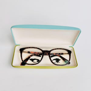 May include: Tortoiseshell pattern eyeglasses in a light blue and green glasses case. The glasses have a rectangular frame with gold-coloured accents. The case is open, revealing the glasses and a white interior.