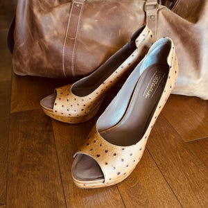 Puede incluir: Un par de tacones de plataforma peep-toe color canela con un patrón de puntos, junto a una bolsa de lona de cuero marrón. Los tacones tienen una plataforma de madera y un interior marrón. La bolsa tiene un asa superior.