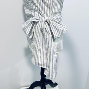 May include: A white and blue striped top with a wrap-around design and a large bow. The top is displayed on a black mannequin, showcasing its style and fit. The fabric has vertical stripes.