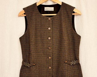 May include: A brown and black houndstooth vest with a black lining and gold buttons. The vest has decorative metal accents near the pockets and the label reads "NORA-YERS". The vest is hanging on a wooden hanger.