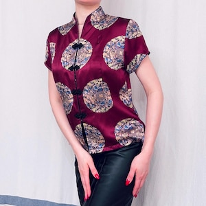 May include: A burgundy satin shirt with a mandarin collar and short sleeves. The shirt features a repeating circular pattern in shades of blue, gold, and cream. Black frog closures and trim accent the front and sleeves. The shirt is paired with black trousers.