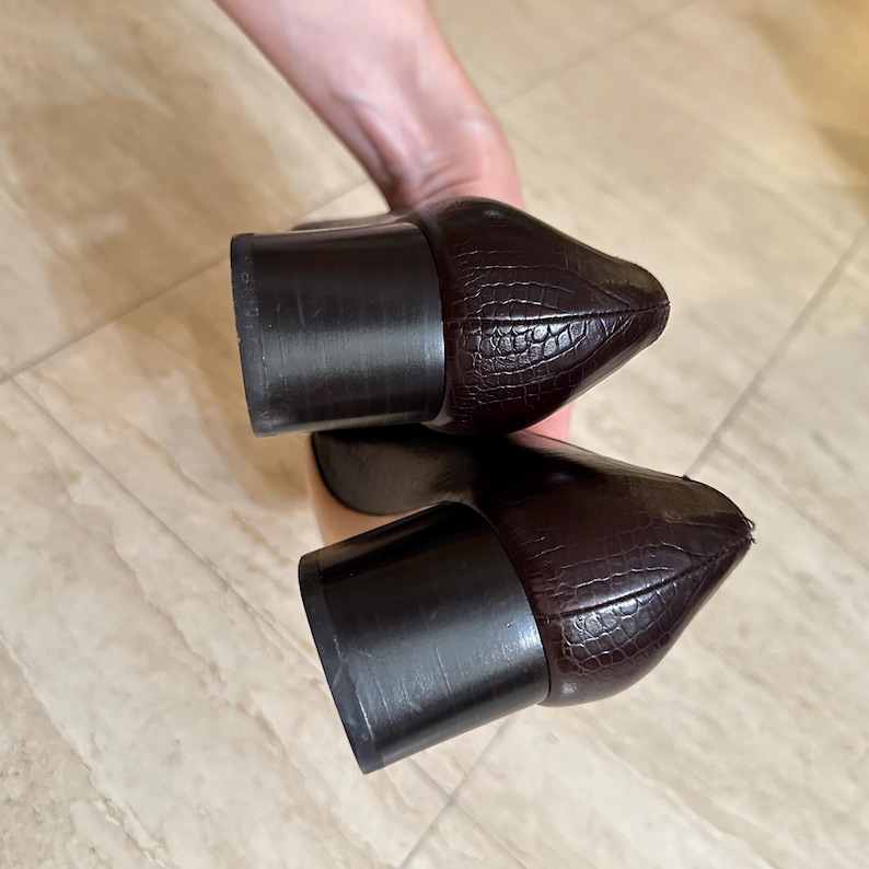 May include: A pair of dark brown, pointed-toe shoes with a textured, crocodile-like pattern. The shoes have block heels. The image is a close-up of the shoes, showcasing their design and shape.